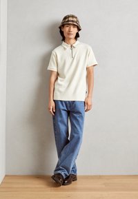 Cream short-sleeve polo with zip collar, textured diagonal pattern. Paired with blue jeans and brown shoes, complemented by a plaid bucket hat.