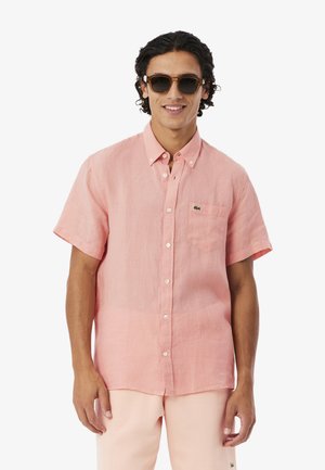 Uomo che indossa una camicia rosa a maniche corte con taschino sul petto e pantaloni beige, indossa anche occhiali da sole, sorridente su sfondo bianco.