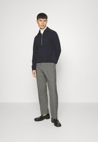 Marineblauer Strickpullover mit halbem Reißverschlusskragen; graue maßgeschneiderte Hose; schwarze Schuhe; das Modell steht vor einem schlichten Hintergrund.