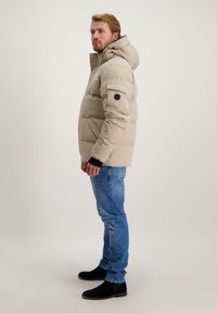 Beige gewatteerde jas met een capuchon, met een rits aan de voorkant, zijzakken en zwarte accenten, gedragen met blauwe jeans en zwarte laarzen.