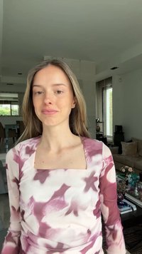 Νέα γυναίκα με μακριά ξανθά μαλλιά που φοράει ένα ροζ και λευκό τοπ με σχέδιο tie-dye, στέκεται σε ένα μοντέρνο σαλόνι με ουδέτερη διακόσμηση.