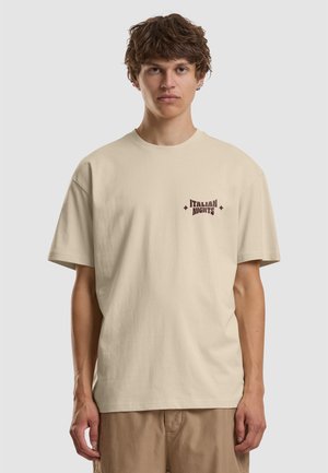 Jeune homme aux cheveux courts et bouclés portant un t-shirt beige "Italian Nights" et un pantalon marron clair sur fond gris uni.
