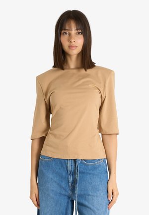 Donna con capelli castani lisci che indossa un top beige con spalle alte e jeans blu, in piedi su uno sfondo bianco semplice.