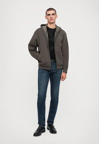 Pelēka zip-up kapuce ar zaļu iekštelpu, valkāta virs tumši melna teksturēta krekla. Saskaņota ar zilām džinsu biksēm un melnām kurpēm.