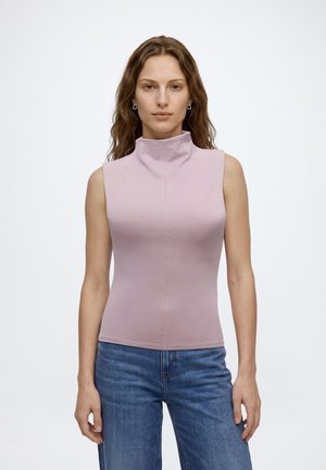 Femme aux cheveux bruns ondulés portant un haut sans manches à col roulé rose clair et un jean bleu, debout devant un fond blanc uni.