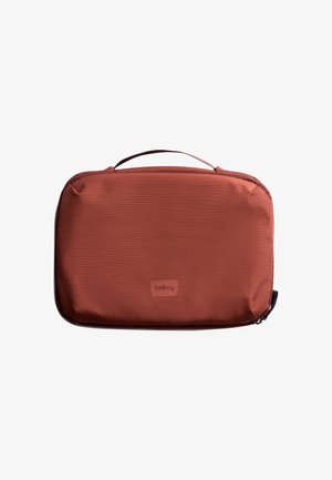 Étui de voyage en tissu rouge avec fermeture éclair, forme rectangulaire plate et poignée supérieure. Présente une texture subtile et le logo de la marque à l'avant.