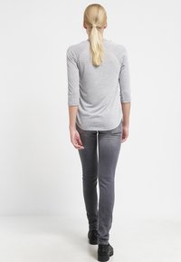 Graues Oberteil mit drei Viertelärmeln, taillierte Passform, glatte Textur, kombiniert mit grauen schmalen Jeans und schwarzen Schuhen mit dicker Sohle, von hinten betrachtet.