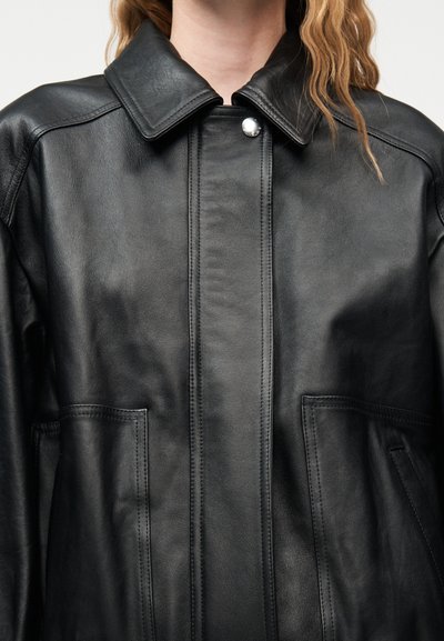 Κοντινό πλάνο ενός ατόμου που φοράει μαύρο δερμάτινο μπουφάν με μυτερό γιακά και κλείσιμο με κουμπιά snap στον λαιμό.