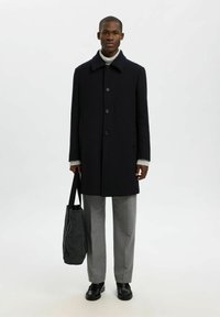 Vīrietis ģērbts melnā pogātā mētelī, pelēkā turtleneck džemperī, pelēkās biksēs, melnās kurpēs, tur rokās melnu tote somu, stāv pret vienkāršu fona.