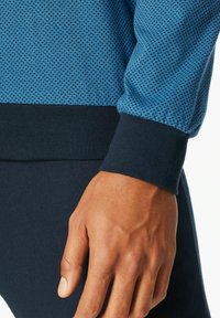 Niebieski sweter z wzorem o teksturowanym wykończeniu, z ciemnym ściągaczem. Zbliżenie przedstawiające dłoń opartą na materiale.