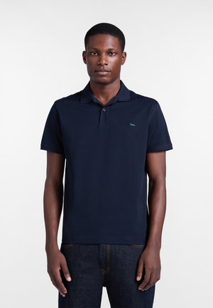 Modelo maschile che indossa una polo a maniche corte blu scuro con un piccolo logo verde ricamato sul petto, abbinata a jeans scuri.