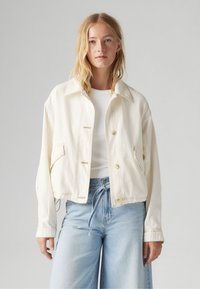 Crème kleurige cropped denim jacket met een spitse kraag, knoopsluiting aan de voorkant en twee zijzakken. Lichtblauwe wijde jeans, met een plain witte top eronder.