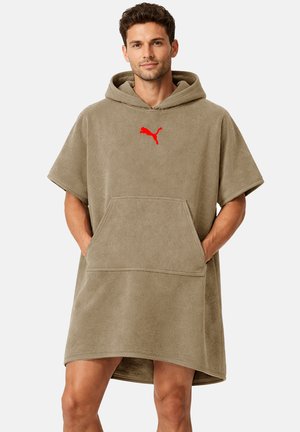 Mann trägt einen khakifarbenen Kapuzen-Handtuchponcho mit rotem Puma-Logo, Hände in der Vordertasche, steht vor einem einfarbigen Hintergrund.