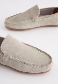 Paire de mocassins en daim beige avec coutures visibles cousues à la main et semelles plates marron sur fond blanc.