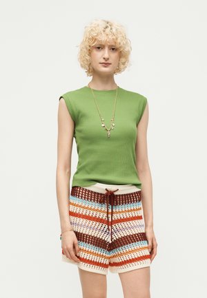 Personne aux cheveux blonds courts et bouclés portant un haut vert sans manches, un short en tricot rayé coloré et un collier en or avec des breloques.