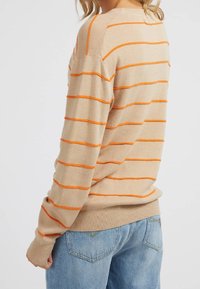 Beige trui met horizontale oranje strepen, geribbelde manchetten en zoom, gemaakt van zacht stof. Draagt met blauwe spijkerbroeken.