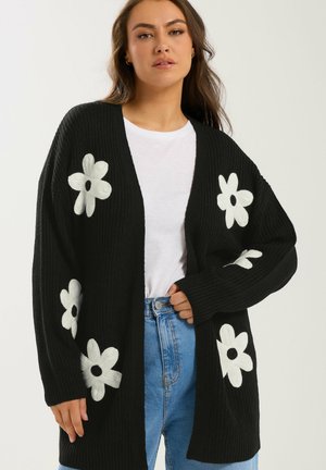 Mujer con cárdigan negro de punto con patrones de flores blancas, camiseta blanca y vaqueros azules, de pie frente a un fondo liso.