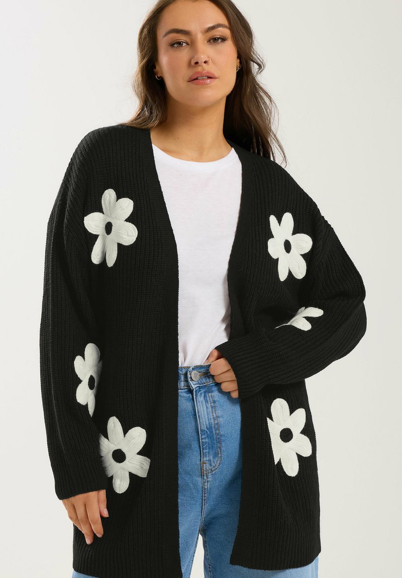 Frau trägt eine schwarze Strickjacke mit weißen Blumenmustern, ein weißes T-Shirt und blaue Jeans, steht vor einem einfarbigen Hintergrund.