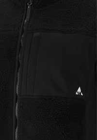 Giacca in pile nera con pannelli lisci, tessuto a pelo alto texturizzato, chiusura con zip e un accento di logo bianco sulla tasca del petto.