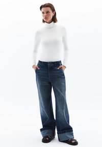 Weißes Rollkragenoberteil, übergroße blaue Jeans mit umgeschlagenen Säumen und schwarze Sandalen. Glatte Textur mit lässiger, lockerer Passform.