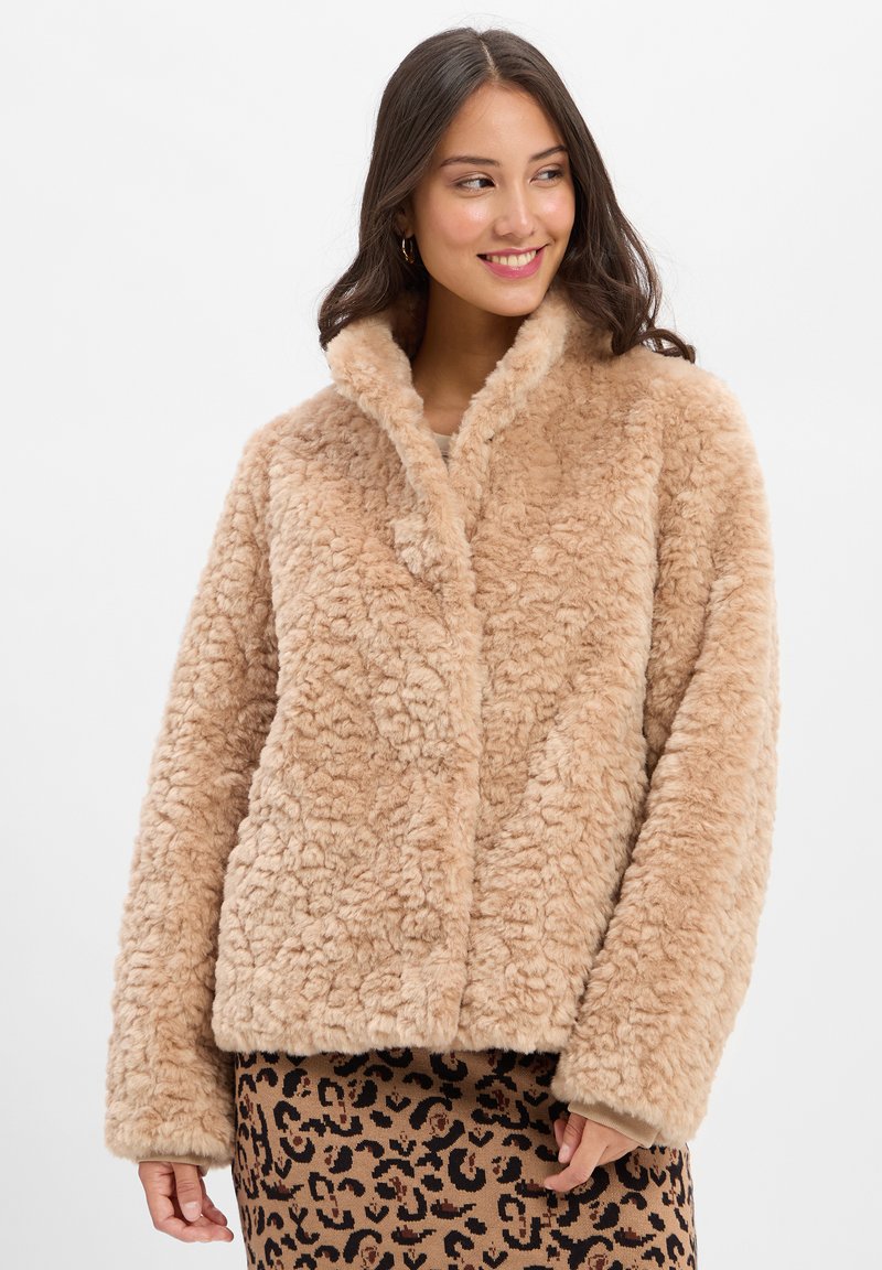 Kurtka z imitacji futra w jasnym beżu z teksturowaną, puszystą powierzchnią, szerokimi klapami i prostym krojem. Połączona z brązową spódnicą w wzór lamparta.