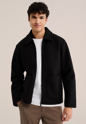 Jeune homme aux cheveux bouclés portant une veste noire zippée, un t-shirt blanc et un pantalon marron, debout avec une main dans la poche.