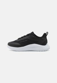 Scarpe da ginnastica nere con una tomaia sintetica liscia, suola in gomma bianca, sistema di allacciatura e un logo discreto di lato. Dettagli testurizzati presenti.