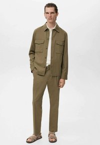 Chaqueta verde oliva de botones con dos bolsillos en el pecho, combinada con pantalones a juego. Lleva puesta una camiseta blanca de canalé y sandalias. Textura suave.