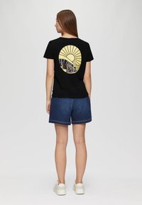 Zwart t-shirt met een cirkelvormige gele grafiek met zonnestralen en de tekst "LAAT DE ZON IN JE HART SCHIJNEN" op de achterkant, gecombineerd met denim shorts.