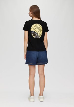 T-shirt noir avec un graphique circulaire jaune comportant des rayons de soleil et le texte "LAISSE LE SOLEIL ENTRER DANS TON COEUR" au dos, associé à un short en denim.
