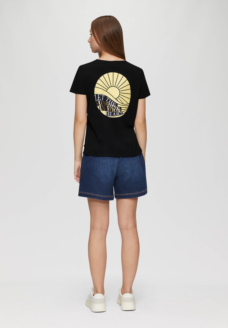 Zwart t-shirt met een cirkelvormige gele grafiek met zonnestralen en de tekst "LAAT DE ZON IN JE HART SCHIJNEN" op de achterkant, gecombineerd met denim shorts.