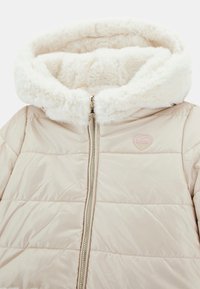 Beżowa kurtka ocieplana z kapturem wyłożonym futrem, z złotym zamkiem błyskawicznym i różowym logo w kształcie serca. Powierzchnia zewnętrzna ma gładką teksturę i pikowaną strukturę.