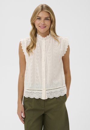 Blondína s úsmevom, oblečená v bielom čipkovanom top bez rukávov so zvlnenými okrajmi a olivovozelených nohaviciach, stojaca pred jednoduchým pozadím.