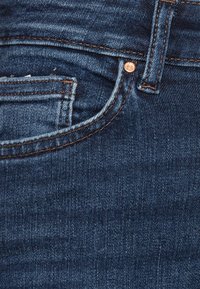 Tmavě modrý denimu s hladkým povrchem, který má malou přední kapsu a měděný knoflík, zvýrazněný kontrastním šitím.