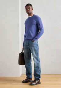 Lila Baumwollpullover mit einem weißen Rundhalsausschnitt, kombiniert mit hellblauen, distressed Jeans. Braune Lederschuhe und eine braune Tasche runden das Outfit ab.