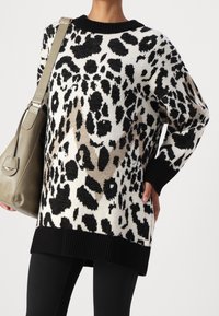 Pull en imprimé léopard noir et blanc avec un col et un ourlet noir côtelés, fabriqué en maille, associé à un sac à main vert et un legging noir.