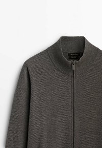 Grauer Pullover mit Reißverschluss, strukturiertem Strickdesign, hohem Kragen und gerippten Bündchen. Verfügt über einen passenden Reißverschluss und ein Etikett am Hals.