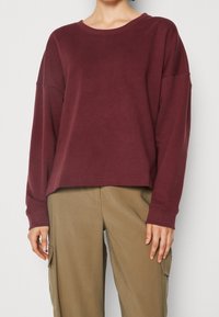 Bordeauxroter Pullover mit langen Ärmeln aus weicher Baumwolle, ausgestattet mit einem breiten Rundhalsausschnitt und entspanntem Schnitt sowie gerippten Bündchen und Saum. Kombiniert mit beigen Hosen.