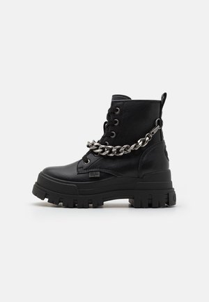Bottines noires en cuir texturé, avec une semelle épaisse, un détail de chaîne proéminent sur le côté et des lacets ronds.