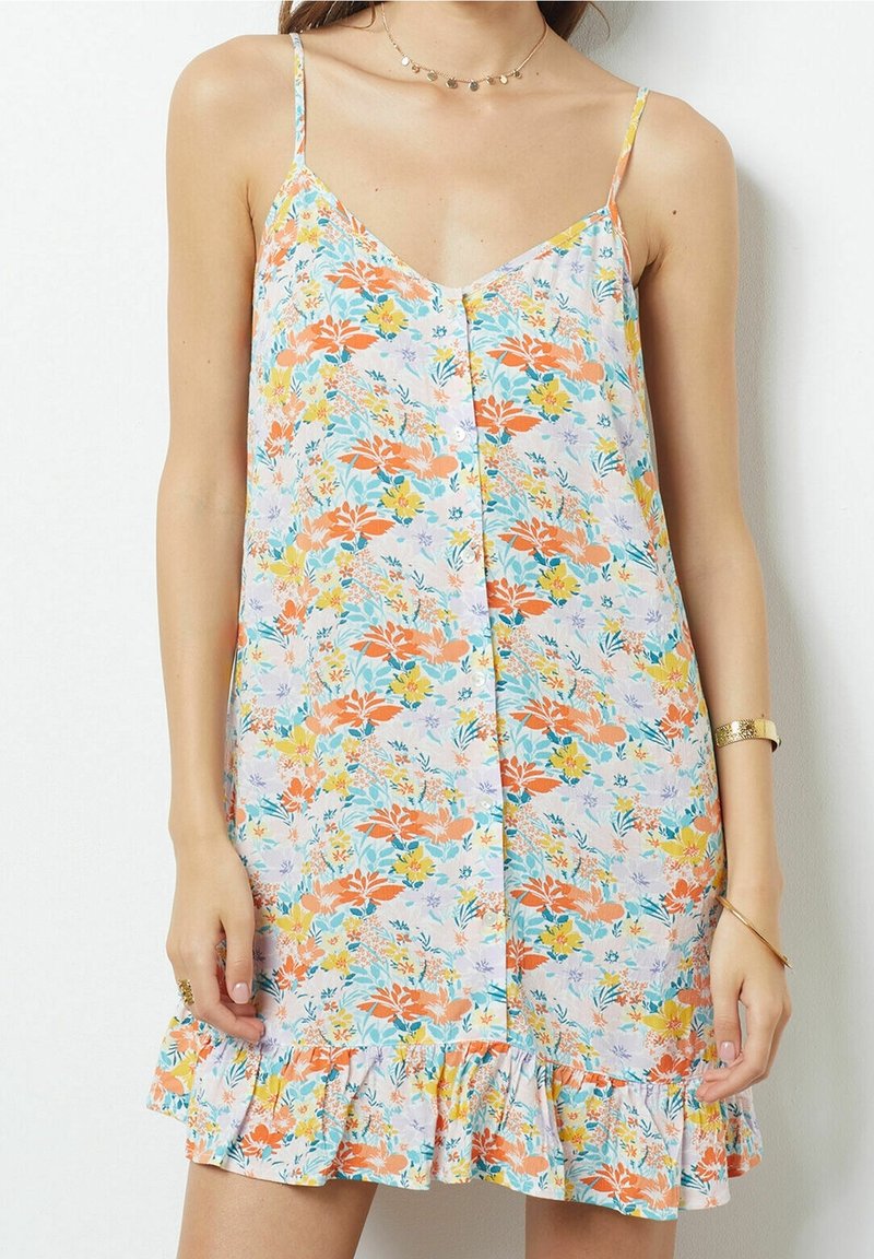 Femme portant une robe sans manches à fleurs orange, jaunes et bleues, avec des boutons sur le devant et un ourlet à volants.