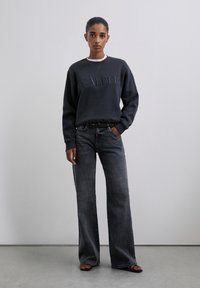 Mujer de pie con un sudadera oscura con el texto "SCALPER", jeans de pierna ancha negros desgastados y sandalias negras de dedo al aire, contra un fondo liso.