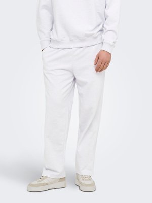 Bărbat purtând pantaloni de trening albi, bluză albă și adidași bej și albi, stând în fața unui fundal simplu.