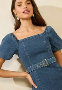 Modra denim obleka s kratkimi napihnjenimi rokavi, kvadratnim izrezom in pasom, ki ga krasi pravokotna zlat zgib. Gladka tekstura.