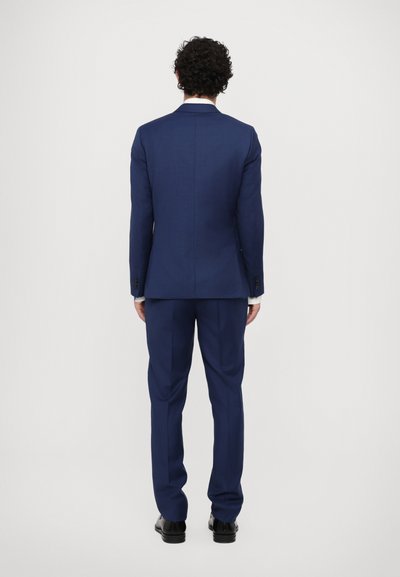 Costume sur mesure bleu marine, avec une fente unique, des revers crantés et un pantalon assorti. Le tissu semble lisse avec une texture subtile.