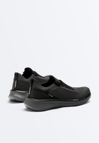 Paar schwarze, flach geschnittene Laufschuhe mit atmungsaktivem Mesh, dicken gepolsterten Sohlen und Schlaufen zum Anziehen auf weißem Hintergrund.