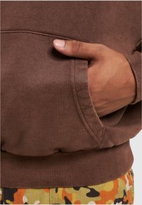 Suéter marrón con textura acanalada y cinturilla ancha, que cuenta con un bolsillo lateral. Los pantalones tienen un patrón de camuflaje multicolor.