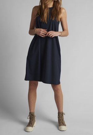 Frau steht mit verschränkten Händen, trägt ein ärmelloses, knieumspielendes navyblaues Kleid und beige Schnürstiefel vor einem neutralen Hintergrund.