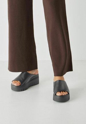 Sandali platform neri con fascia larga e punta aperta. Suola rialzata con battistrada texturizzato. Abbinati a pantaloni marroni a gamba larga.