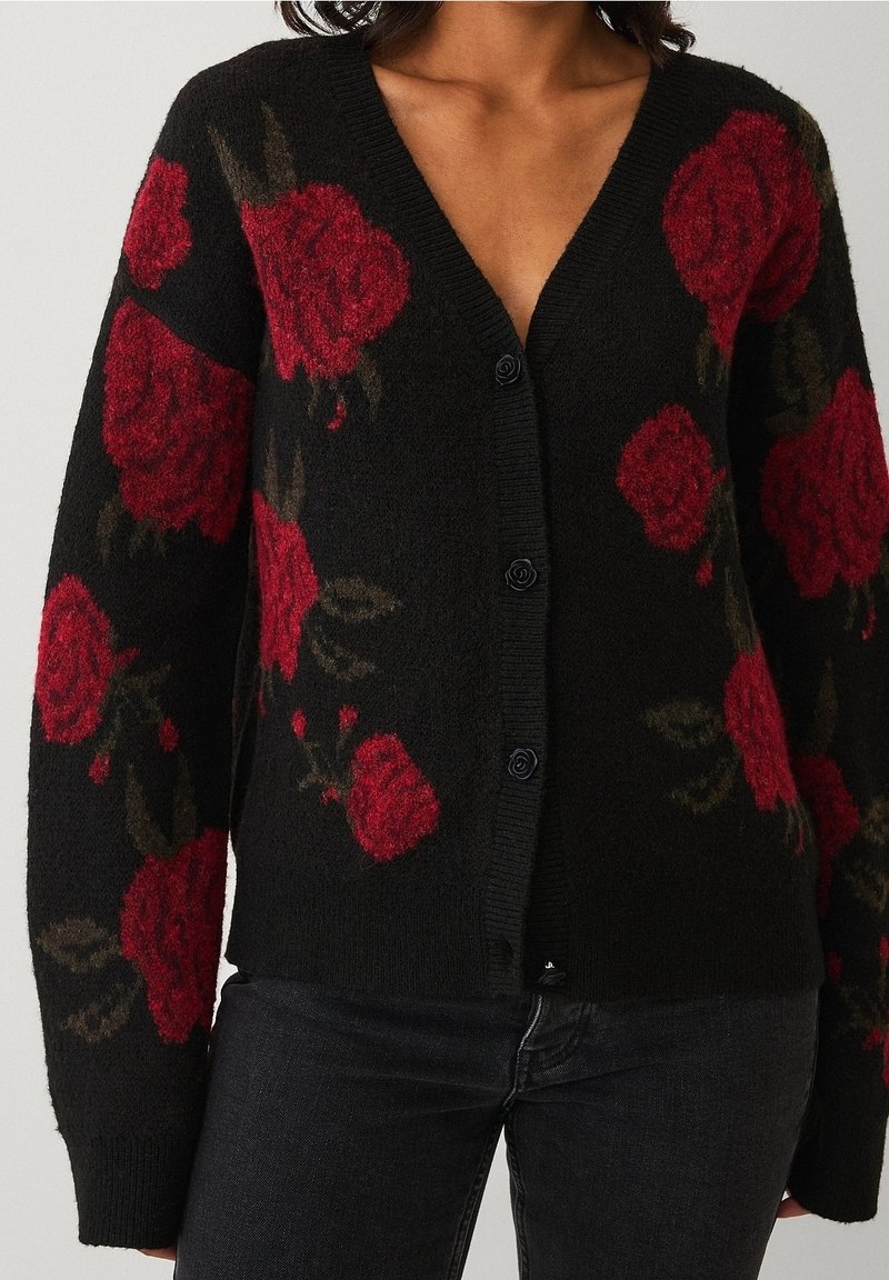 Cardigan noir boutonné avec de grands motifs de roses rouges, porté sur un pantalon noir, montré de la poitrine aux hanches.
