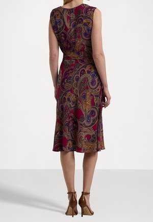 Femme debout portant une robe sans manches mi-longue à motif cachemire bordeaux, bleu et or avec des sandales marron à talons hauts, vue de dos.
